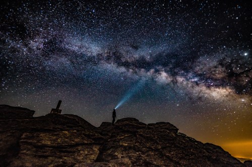 A silhouette of a person on rocks, shining a flashlight at a starry sky with the Milky Way visible.
