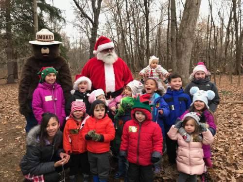 A group of children in winter clothing poses with Santa and a bear mascot in a wooded area.
