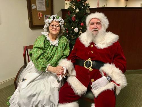 A man in a Santa suit and a woman in a green dress with a white apron sit together by a Christmas tree.