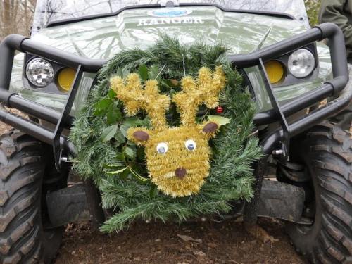 A festive wreath with a reindeer face decorates the front of an all-terrain vehicle in a wooded area.