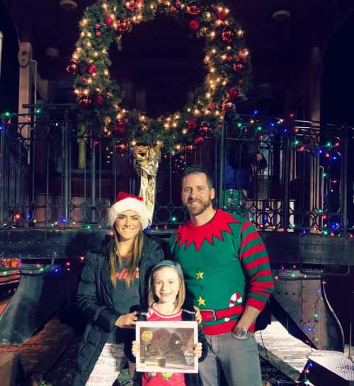 A family poses with a holiday wreath and lights, holding a framed award, dressed in festive attire.