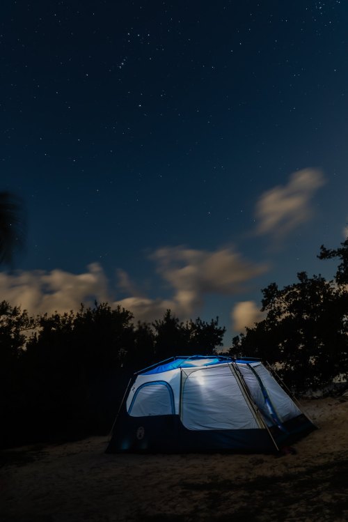 A tent illuminated at night under a starry sky, surrounded by trees and soft clouds.