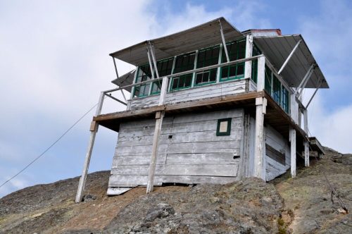 A wooden lookout tower perched on a rocky hillside, featuring large green windows and a flat roof under a cloudy sky.