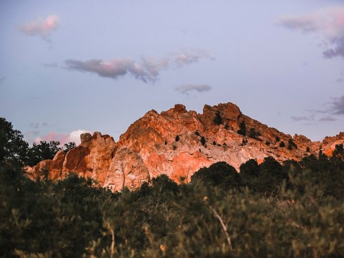 Sunset over rugged red rock formations, surrounded by green foliage and a pastel sky with scattered clouds.