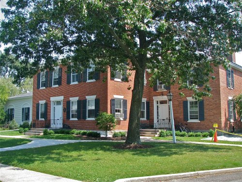 A brick house with white doors and black shutters, surrounded by green grass and trees on a sunny day.