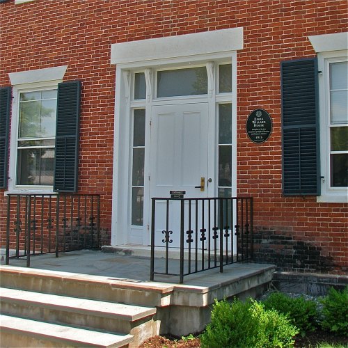Historic building entrance with white door, black shutters, and a plaque on the brick wall. Steps lead to the door.