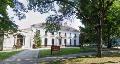 A white building with a large entrance, surrounded by trees and a grassy area, under a clear sky.