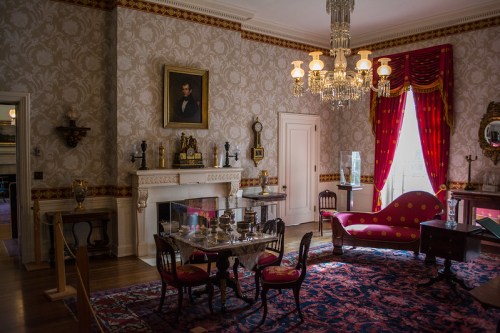 A vintage room with ornate wallpaper, a chandelier, a red sofa, and a dining table, featuring elegant decor and artwork.