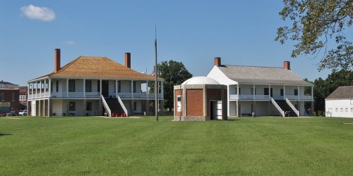 Two historic buildings with porches and gabled roofs, set on a grassy field under a clear blue sky.