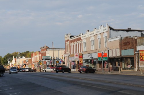 A small town street lined with historic buildings, shops, and parked cars under a cloudy sky.