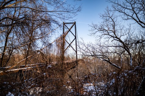 A snow-covered landscape featuring a suspension bridge framed by bare trees against a clear blue sky.