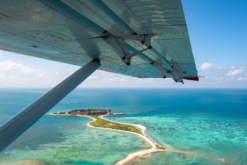 Aerial view from a plane wing over a tropical island surrounded by turquoise waters and coral reefs.