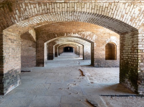 A long, empty corridor with arched brick ceilings and walls, featuring a sandy floor and dim lighting.