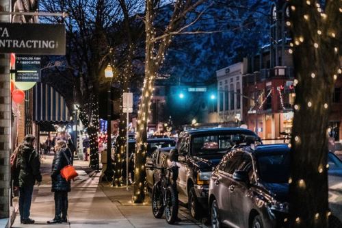 A bustling street at night, lined with trees adorned with lights, cars parked, and people chatting near a storefront.