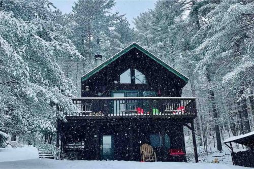 A cozy black cabin surrounded by snow-covered trees, with a balcony and colorful chairs, in a winter landscape.