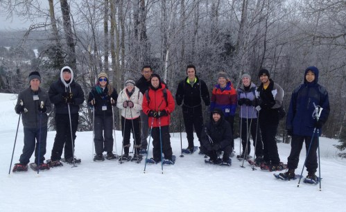 A group of 12 people in winter gear stand on snow, surrounded by trees, smiling for a photo during a snowshoeing activity.