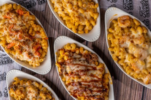 A variety of macaroni and cheese dishes topped with different ingredients, served in white bowls on a wooden table.