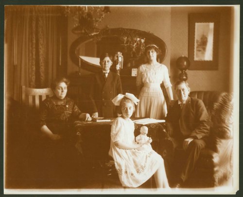 A sepia-toned photo of five people, including a girl with a doll, posing in a vintage living room setting.