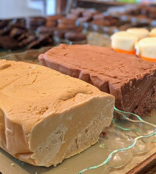 Two blocks of fudge, one light beige and one brown, displayed on a glass platter with assorted cookies in the background.