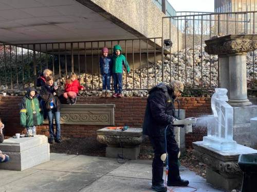 A person carves an ice sculpture while children watch from a nearby ledge in a park setting.