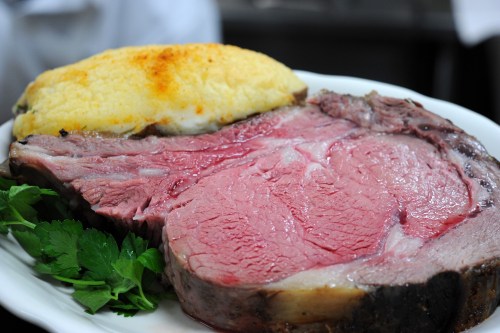 A plate featuring a large slice of pink prime rib with a golden-brown topping and garnished with fresh herbs.