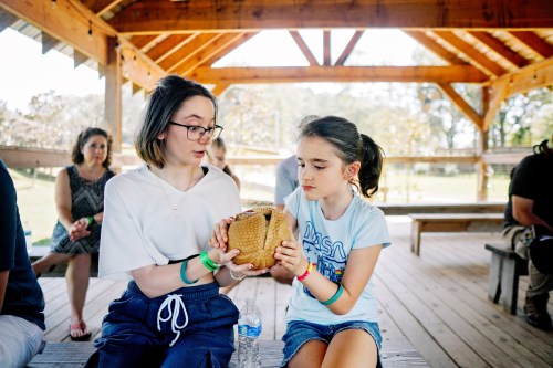 A woman and a girl share a large, round object while sitting in a wooden pavilion, with others in the background.