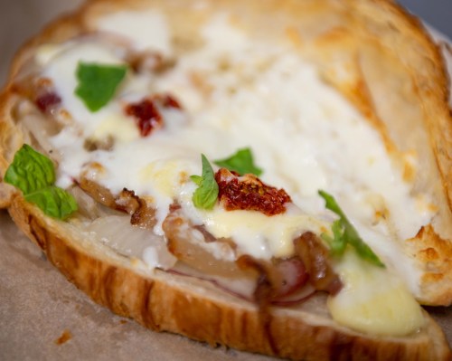A close-up of a toasted sandwich with melted cheese, caramelized onions, sun-dried tomatoes, and fresh herbs.