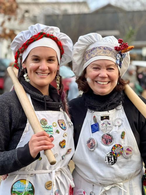 Two women in chef hats and aprons, smiling and holding wooden rolling pins, adorned with various badges and pins.