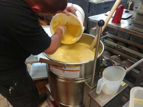 A person pours a large bucket of yellow soup into a stainless steel pot in a commercial kitchen.