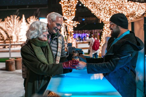 A smiling elderly couple receives a warm drink from a vendor at a festive, illuminated outdoor event.