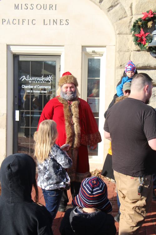Santa Claus stands outside a building, surrounded by children and adults, with festive decorations in the background.