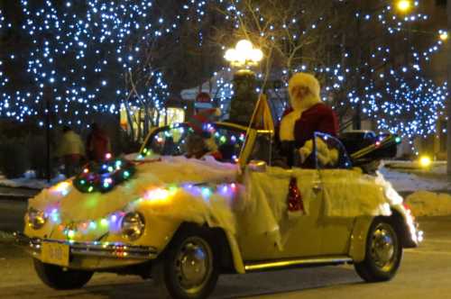 A festive convertible car decorated with lights and snow, featuring Santa Claus waving from the driver's seat.