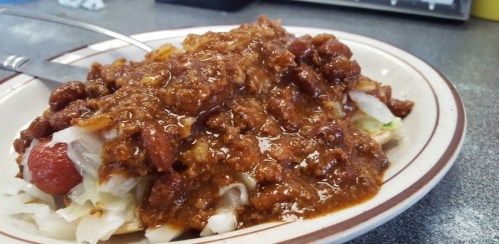 A plate of chili topped with beans over shredded lettuce and a hot dog, served with a fork on a gray table.