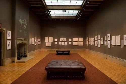 A spacious art gallery with framed artworks on the walls and two brown leather benches in the center.