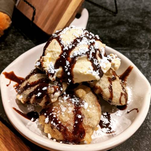 A bowl of dessert featuring fried dough balls topped with ice cream, chocolate sauce, and powdered sugar.