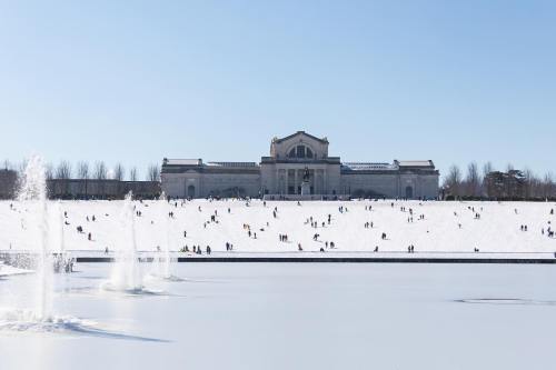 A snowy landscape with people playing in the snow, featuring a large building in the background and a frozen pond in the foreground.