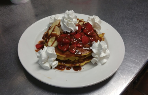 A stack of pancakes topped with strawberries, chocolate sauce, and whipped cream, served on a white plate.