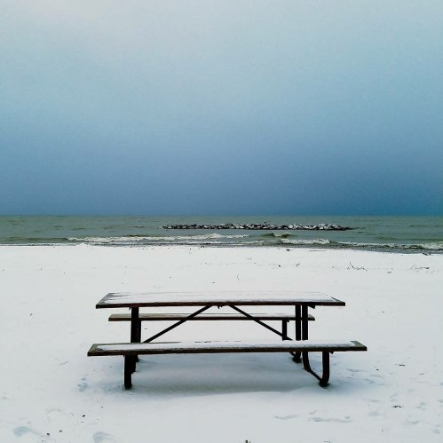 A snow-covered picnic table on a beach, with a gray sky and calm water in the background.
