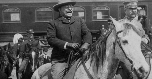 Historical black-and-white photo of a man on horseback, smiling, with soldiers and a train in the background.
