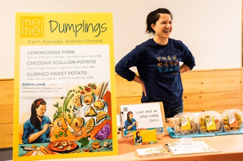 A smiling woman stands behind a colorful dumpling display, showcasing various flavors and cooking classes.