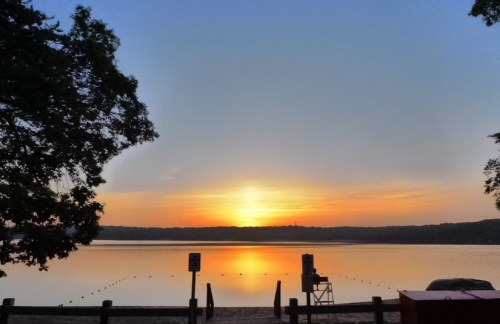 A serene lake at sunset, with silhouettes of trees and a lifeguard chair in the foreground.