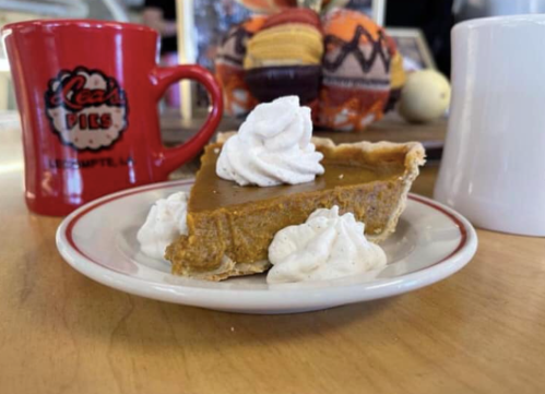A slice of pumpkin pie topped with whipped cream, served on a plate with red and white mugs in the background.