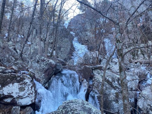 A frozen waterfall cascades down rocky terrain, surrounded by bare trees and rugged boulders.