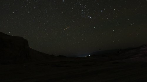 A starry night sky filled with stars and constellations, with a faint shooting star visible above a dark landscape.