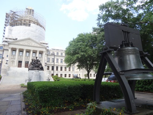 A large bell stands in front of a historic building with a dome under construction, surrounded by greenery.