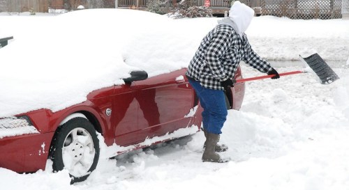 A person in a hooded jacket shovels snow off a red car buried in a snowy driveway.