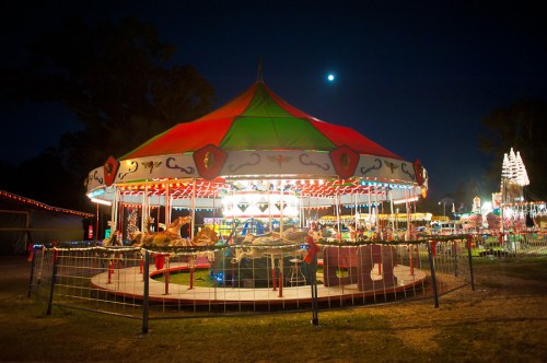 A brightly lit carousel at night, surrounded by festive lights and decorations, with a full moon in the sky.
