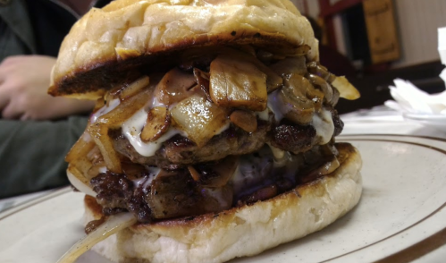 A towering burger with two patties, sautéed onions, and creamy sauce, served on a toasted bun.