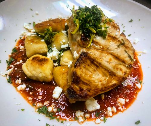 Grilled fish fillet atop a bed of gnocchi in tomato sauce, garnished with herbs and lemon zest.