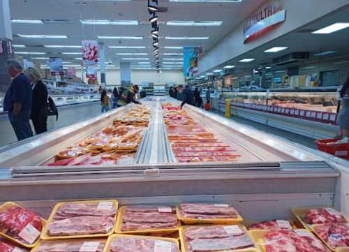 A grocery store meat section with various packaged meats displayed in freezers, shoppers browsing in the background.